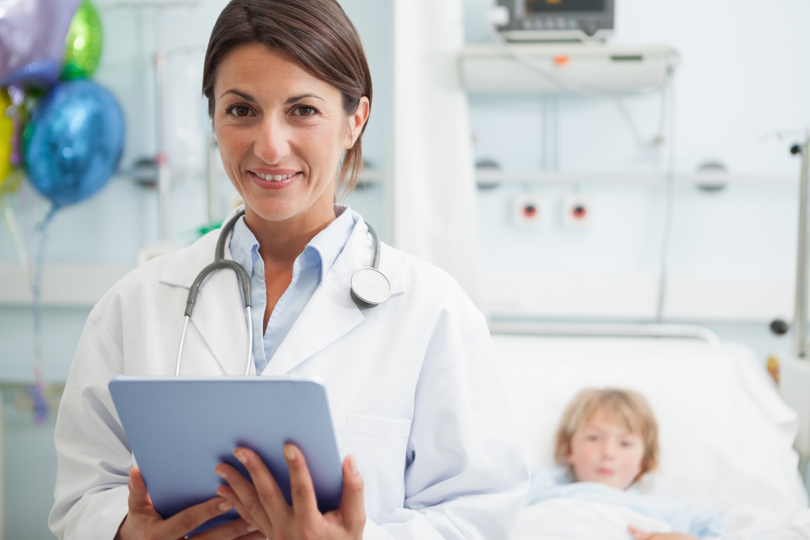Doctor at work in a patient's room holding a Tablet and smiling at the camera. A child patient is on the bed in the background on the right-hand side. There are balloons in the background on the other side of the doctor.
