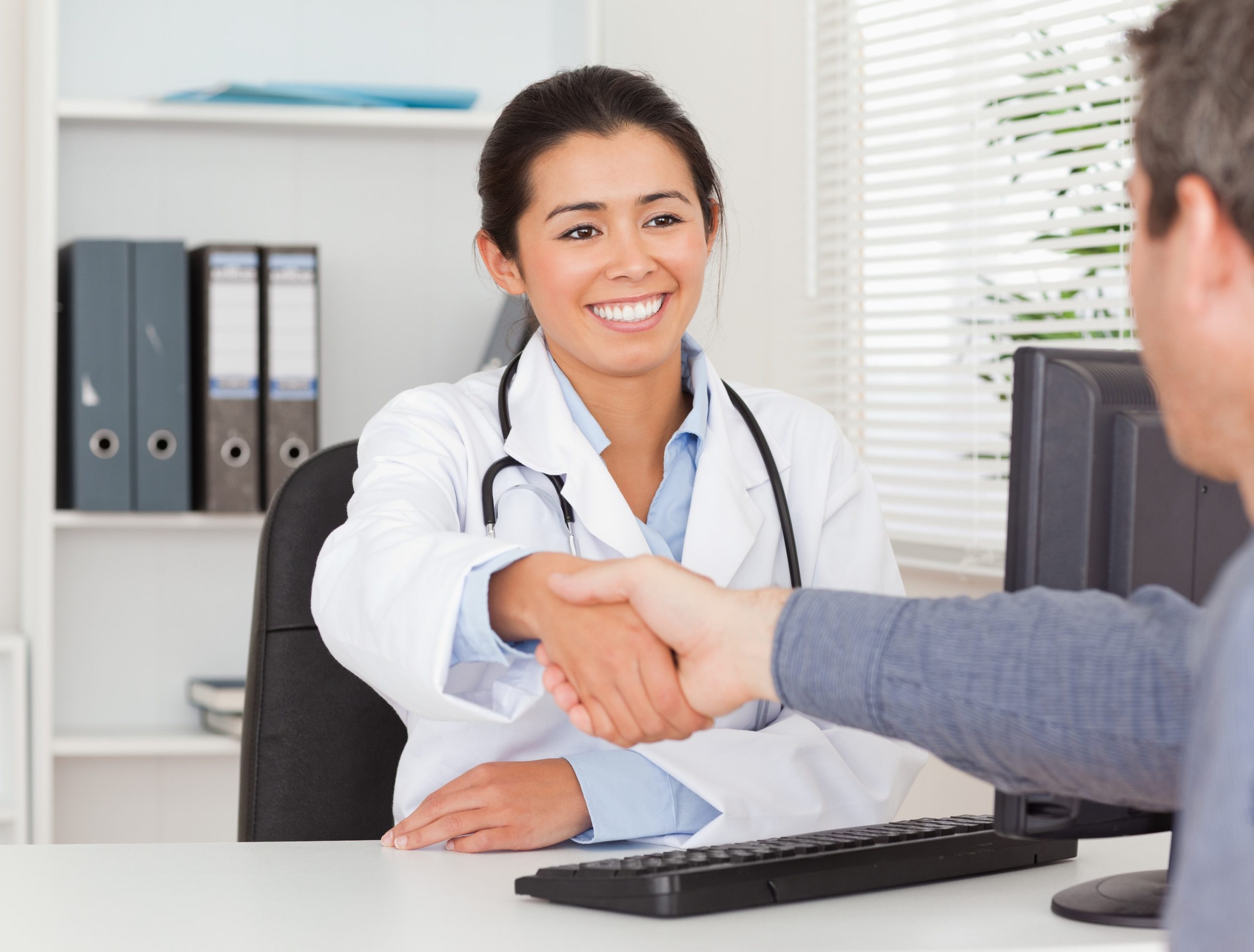 Doctor smiling and shaking hands with a presumed patient. They are both sitting across from each other at a desk.